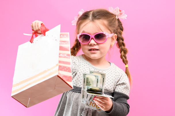 Kleines Mädchen mit Brille, Päckchen und Geld auf rosa Hintergrund, Kind und Einkauf. neu - Foto, Bild