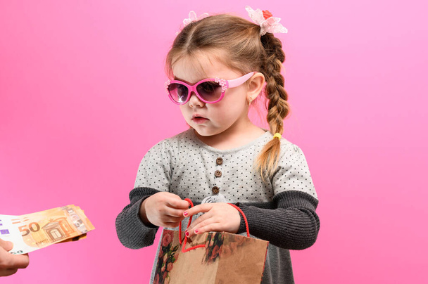 Little cute girl na różowym tle z torbą i pieniędzy, temat zakupów z dziećmi. nowe - Zdjęcie, obraz
