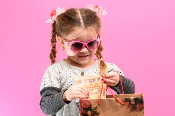 Little cute girl na różowym tle z torbą i pieniędzy, temat zakupów z dziećmi. nowe - Zdjęcie, obraz