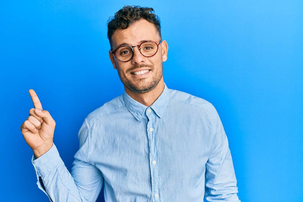 Fiatal spanyol férfi alkalmi ruhát és szemüveget visel, nagy mosollyal az arcán, mutatóujjával a kamerába néz..  - Fotó, kép