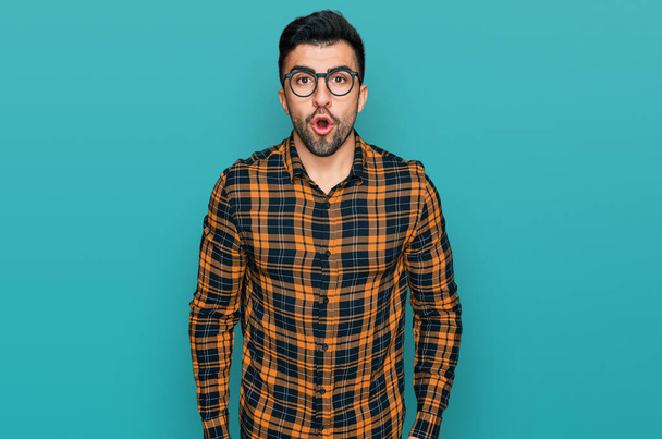 Homme hispanique avec barbe portant des vêtements décontractés et des lunettes effrayé et étonné avec la bouche ouverte pour la surprise, visage incrédule  - Photo, image