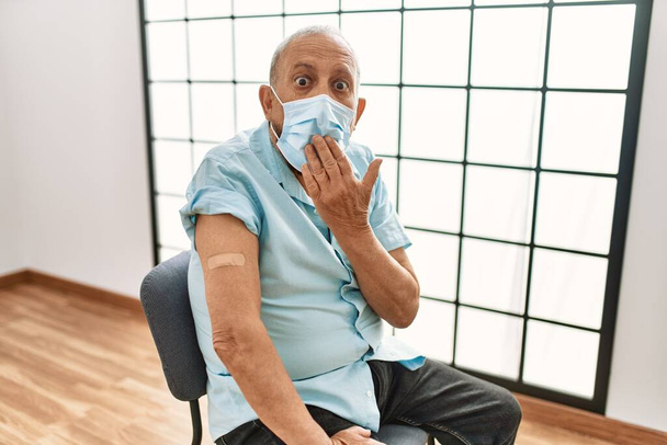 Mature hispanic man with grey hair getting vaccinated covering mouth with hand, shocked and afraid for mistake. surprised expression  - Photo, Image