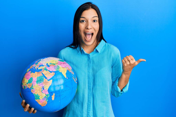 Joven latina sosteniendo gran bola del mundo apuntando el pulgar hacia el lado sonriendo feliz con la boca abierta  - Foto, imagen