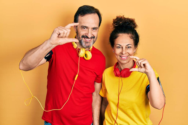 Middle age couple of hispanic woman and man wearing sportswear and arm band smiling and confident gesturing with hand doing small size sign with fingers looking and the camera. measure concept.  - Photo, Image