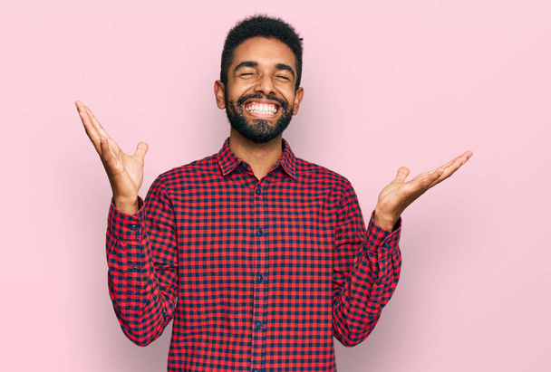 Νεαρός Αφροαμερικάνος που φοράει καθημερινά ρούχα γιορτάζει τρελός και τρελός για την επιτυχία με τα χέρια σηκωμένα και τα κλειστά μάτια να ουρλιάζουν ενθουσιασμένοι. έννοια νικητή  - Φωτογραφία, εικόνα
