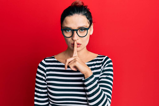 Jeune femme caucasienne portant des vêtements décontractés et des lunettes demandant à se taire avec le doigt sur les lèvres. silence et concept secret.  - Photo, image