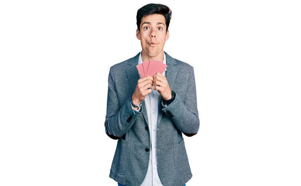 Junger hispanischer Geschäftsmann, der sein Gesicht mit Karten bedeckt, macht Fischgesicht mit Mund und schielenden Augen, verrückt und komisch.  - Foto, Bild