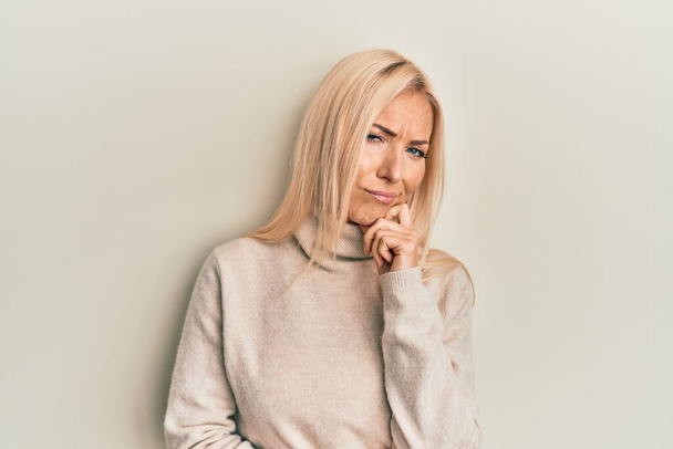 Mujer rubia joven que usa suéter casual de cuello alto de invierno pensando concentrado en la duda con el dedo en la barbilla y mirando hacia arriba preguntándose  - Foto, Imagen