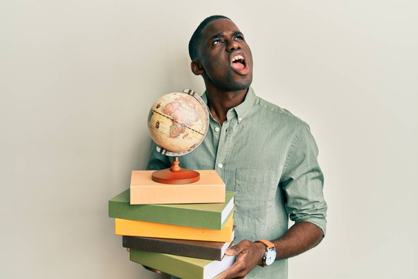 Mladý africký Američan drží vintage světový míč a knihy naštvaný a šílený křičí frustrovaný a zuřivý, křičí hněvem vzhlíží.  - Fotografie, Obrázek