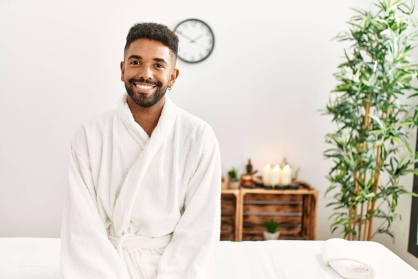Young african american man smiling happy sitting on massage table at beauty center. - 写真・画像