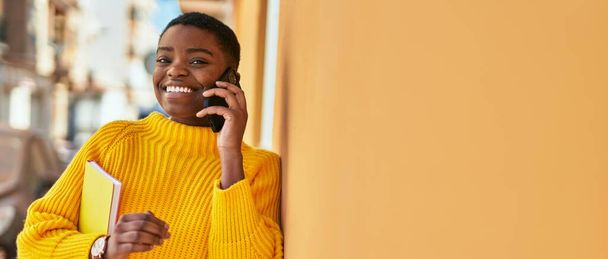 Genç Afrikalı Amerikalı kadın gülümsüyor. Şehirdeki akıllı telefondan mutlu bir şekilde konuşuyor. - Fotoğraf, Görsel