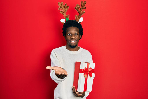 Νεαρός Αφρικάνος Αμερικανός άνδρας φορώντας ελάφι χριστουγεννιάτικο καπέλο κρατώντας δώρο χαμογελώντας χαρούμενα προσφέροντας παλάμη χέρι δίνοντας βοήθεια και αποδοχή.  - Φωτογραφία, εικόνα