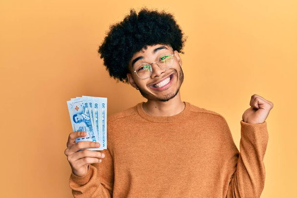 Young african american man with afro hair holding 50 thai baht banknotes screaming proud, celebrating victory and success very excited with raised arm  - Foto, Imagen