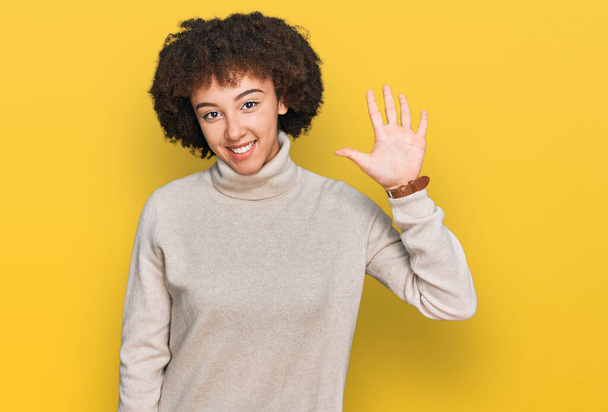Νεαρή ισπανόφωνη κοπέλα που φοράει μάλλινο χειμωνιάτικο πουλόβερ που δείχνει και δείχνει προς τα πάνω με τα δάχτυλα νούμερο πέντε χαμογελώντας σίγουρη και χαρούμενη.  - Φωτογραφία, εικόνα