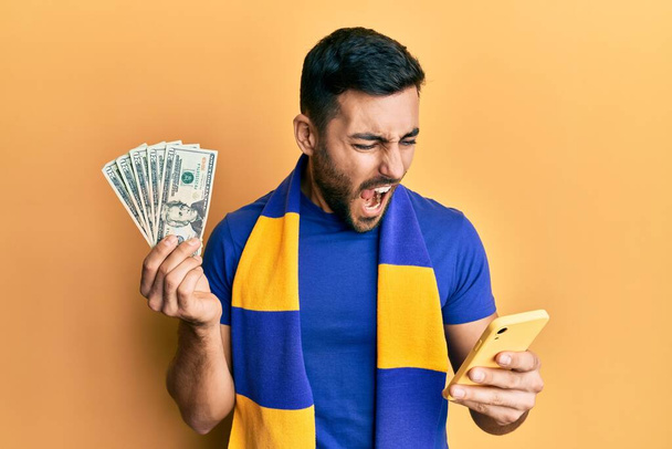Joven partidario del fútbol hispano usando un teléfono inteligente sosteniendo billetes en dólares estadounidenses enojado y loco gritando frustrado y furioso, gritando con ira. rabia y concepto agresivo.  - Foto, imagen