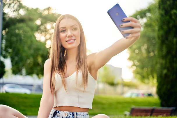Glücklich lächelnde blonde Frau macht Selfie mit dem Handy, im Freien.Konzept des Urlaub Lebensstil. Schöne Mädchen mit Smartphone im Park.Sonniger Tag. Glückskonzept. - Foto, Bild