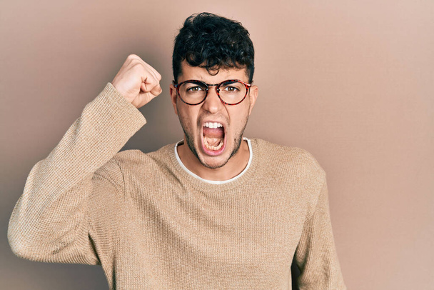 Junger hispanischer Mann in lässiger Kleidung und Brille, wütend und verrückt, hebt frustriert und wütend die Faust und schreit vor Wut. Wut und aggressives Konzept.  - Foto, Bild