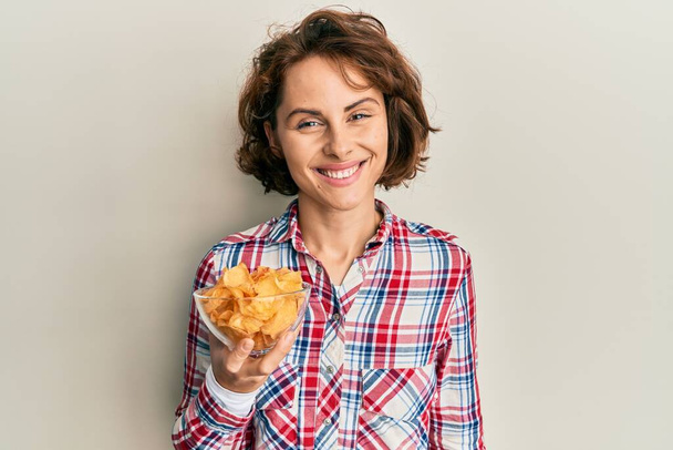 Young brunette woman holding potato chips looking positive and happy standing and smiling with a confident smile showing teeth  - Foto, Imagem
