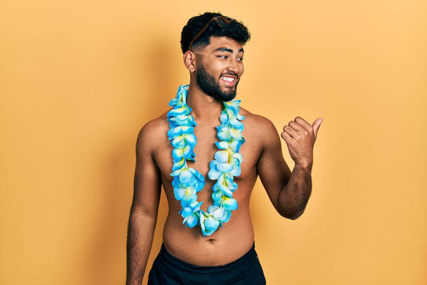 Arabische man met baard in badpak en hawaiiaanse lei glimlachend met vrolijk gezicht en wijzend naar de zijkant met duim omhoog.  - Foto, afbeelding