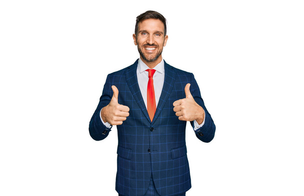 Knappe man met baard in zakenpak en stropdas teken van succes doet positief gebaar met de hand, duimen omhoog glimlachend en gelukkig. vrolijke uitdrukking en winnaar gebaar.  - Foto, afbeelding