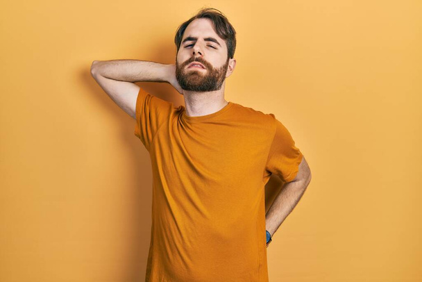 Uomo caucasico con barba che indossa maglietta gialla casual che soffre di dolore al collo, toccando collo con mano, dolore muscolare  - Foto, immagini