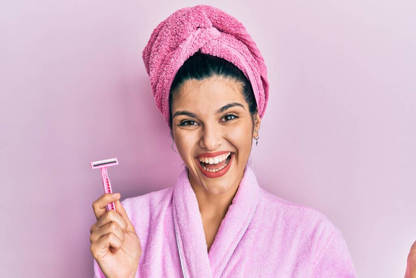Fiatal spanyol nő visel zuhany fürdőköpeny gazdaság borotva ünneplő teljesítmény boldog mosollyal és a győztes kifejezés emelt kézzel  - Fotó, kép