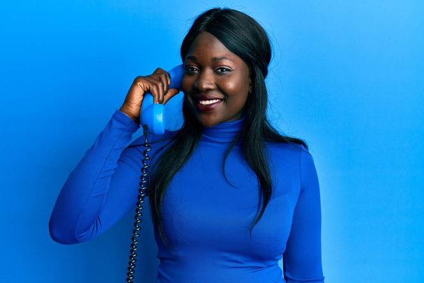Afrikanische junge Frau, die am Telefon spricht, sieht im Stehen positiv und glücklich aus und lächelt mit einem selbstbewussten Lächeln, das Zähne zeigt  - Foto, Bild