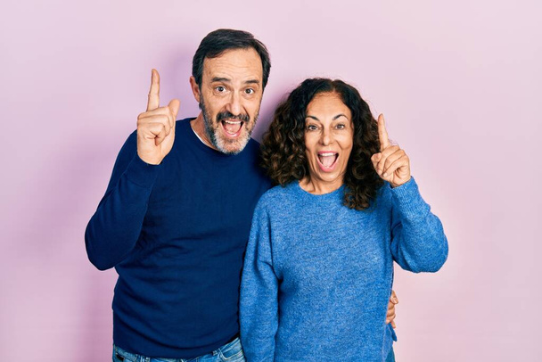 Пара латиноамериканцев среднего возраста обнимаются и стоят вместе улыбаясь удивлённые и удивлённые и указывающие пальцами и поднятыми руками.  - Фото, изображение