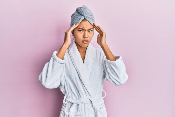 Mujer joven china que usa gorra de toalla de ducha y albornoz con la mano en la cabeza para el dolor en la cabeza porque el estrés. padecer migraña.  - Foto, Imagen