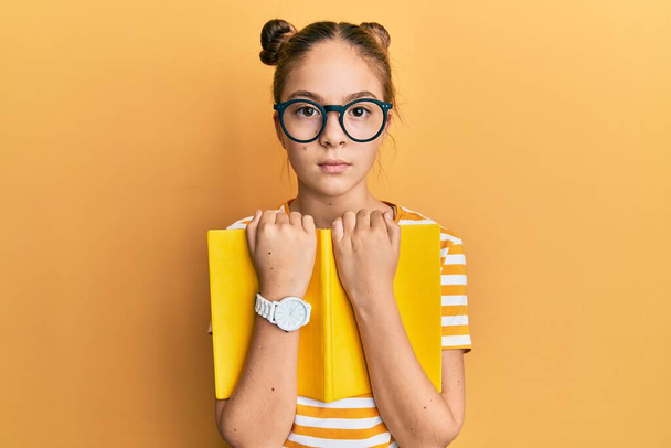 Piękna brunetka dziewczynka nosi okulary i trzyma książkę zrelaksowany z poważnym wyrazem twarzy. proste i naturalne patrzenie w kamerę.  - Zdjęcie, obraz