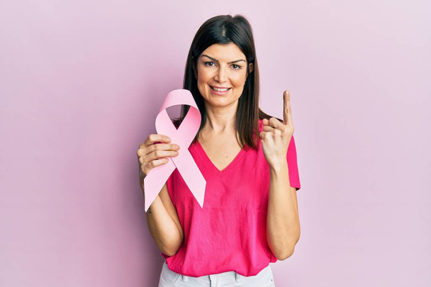 Young hispanic woman holding pink cancer ribbon smiling with an idea or question pointing finger with happy face, number one  - Zdjęcie, obraz