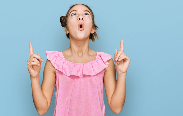 Mooi brunette klein meisje dragen zomer roze shirt verbaasd en verrast omhoog kijken en wijzen met vingers en opgeheven armen.  - Foto, afbeelding