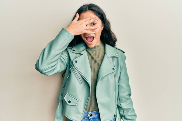 Hispanisches Teenager-Mädchen mit Zahnspange in grüner Lederjacke, das schockiert Gesicht und Augen mit der Hand verdeckt und ängstlich durch die Finger schaut  - Foto, Bild