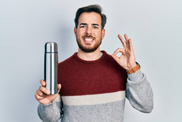Handsome caucasian man with beard holding thermo doing ok sign with fingers, smiling friendly gesturing excellent symbol  - Foto, afbeelding