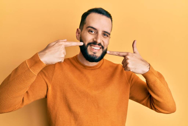 Nuori mies, jolla on parta yllään rento talvi villapaita hymyilevä iloinen osoittaa ja osoittaa sormilla hampaat ja suu. hammasterveyden käsite.  - Valokuva, kuva