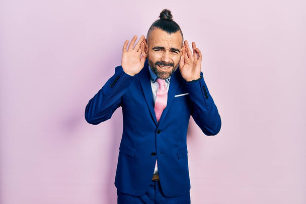 Νεαρός Ισπανός που φοράει κοστούμι και γραβάτα προσπαθώντας να ακούσει και τα δύο χέρια σε μια χειρονομία αυτιού, περίεργος για κουτσομπολιά. πρόβλημα ακοής, κωφός  - Φωτογραφία, εικόνα
