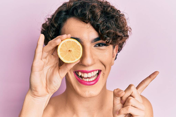 Jongeman draagt vrouw make-up houden citroen glimlachen gelukkig wijzen met hand en vinger naar de zijkant  - Foto, afbeelding