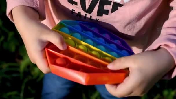 Un niño juega en la calle con un juguete sensorial pop it. La chica presiona coloridas burbujas de silicona suave arco iris suave. Alivio del estrés y la ansiedad. Moda inquieto juego - Imágenes, Vídeo