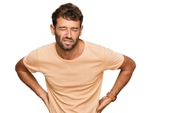 Schöner junger Mann mit Bart trägt lässiges T-Shirt mit Rückenschmerzen, Berührung mit der Hand, muskuläre Schmerzen  - Foto, Bild