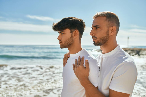 Νεαρό γκέι ζευγάρι με σοβαρή έκφραση που κοιτά στον ορίζοντα στην παραλία. - Φωτογραφία, εικόνα