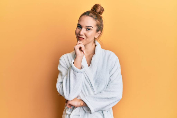 Young blonde woman wearing bathrobe looking confident at the camera with smile with crossed arms and hand raised on chin. thinking positive.  - Foto, imagen