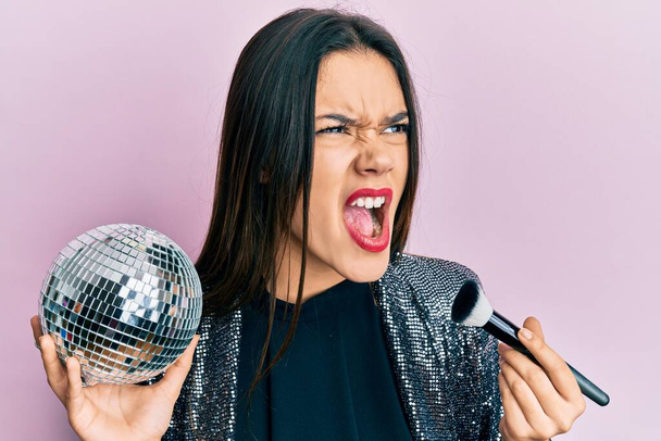 Young hispanic girl holding shiny disco ball and makeup brush angry and mad screaming frustrated and furious, shouting with anger. rage and aggressive concept.  - Fotoğraf, Görsel