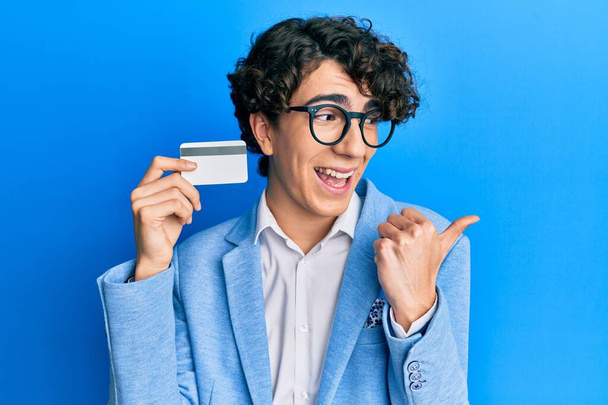 Hispanic young man holding credit card pointing thumb up to the side smiling happy with open mouth  - Фото, зображення