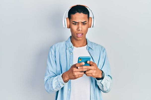Jovem afro-americano usando smartphone usando fones de ouvido assustado e surpreso com a boca aberta para surpresa, rosto descrença  - Foto, Imagem
