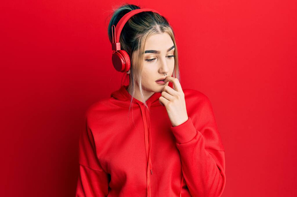 Молода сучасна дівчина носить спортивний одяг і використовує навушники, які виглядають напруженими і нервують руками на роті, кусаючи нігті. проблема тривоги
.  - Фото, зображення