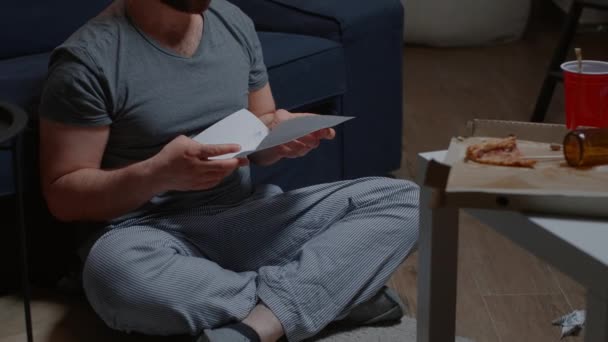 Σοκαρισμένος άνθρωπος που κάθεται στο πάτωμα στο σπίτι διαβάζοντας άσχημα νέα κρατώντας έγγραφα - Πλάνα, βίντεο