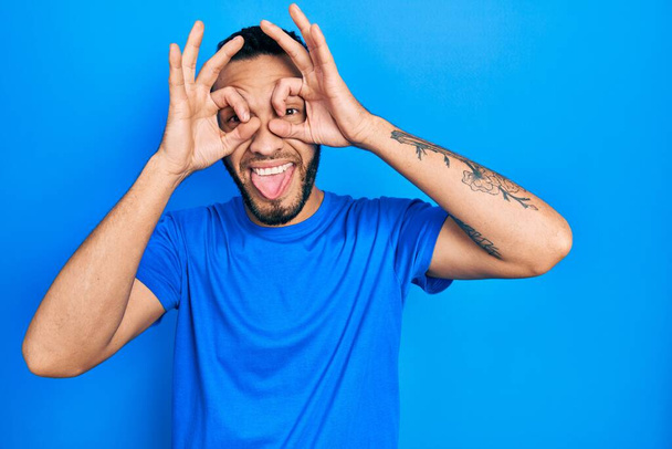 Латиноамериканец с бородой в повседневной синей футболке делает нормальный жест, как бинокль высовывает язык, глаза смотрят сквозь пальцы. сумасшедшее выражение.  - Фото, изображение