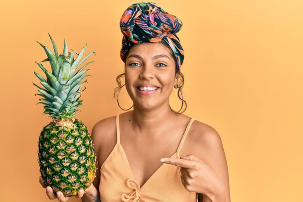 Jeune fille hispanique portant des cheveux turban tenant ananas souriant heureux pointant avec la main et le doigt  - Photo, image