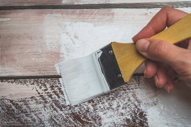 Το γυναικείο χέρι κρατά ένα πινέλο και ζωγραφίζει μια ξύλινη σανίδα σε λευκό χρώμα. Έννοια επισκευής και κατασκευής - Φωτογραφία, εικόνα