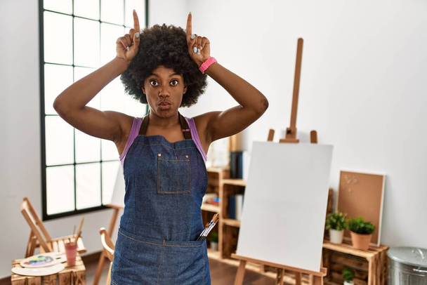 Молодая африканская американка с африканскими волосами в художественной студии делает смешной жест с пальцем над головой, как рога быка  - Фото, изображение